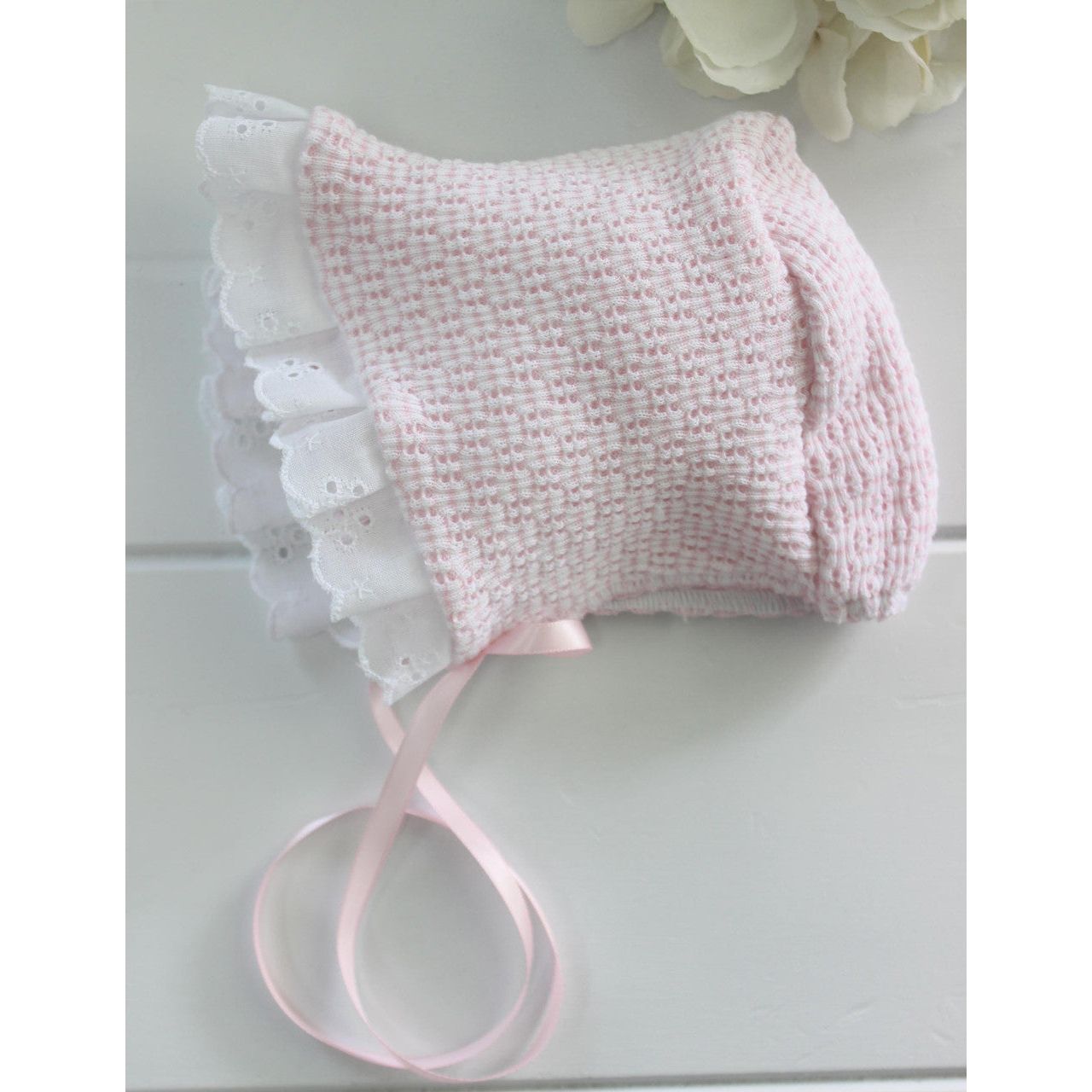 A pink knit bonnet with white eyelet trim and pink satin ribbon ties on a white surface.
